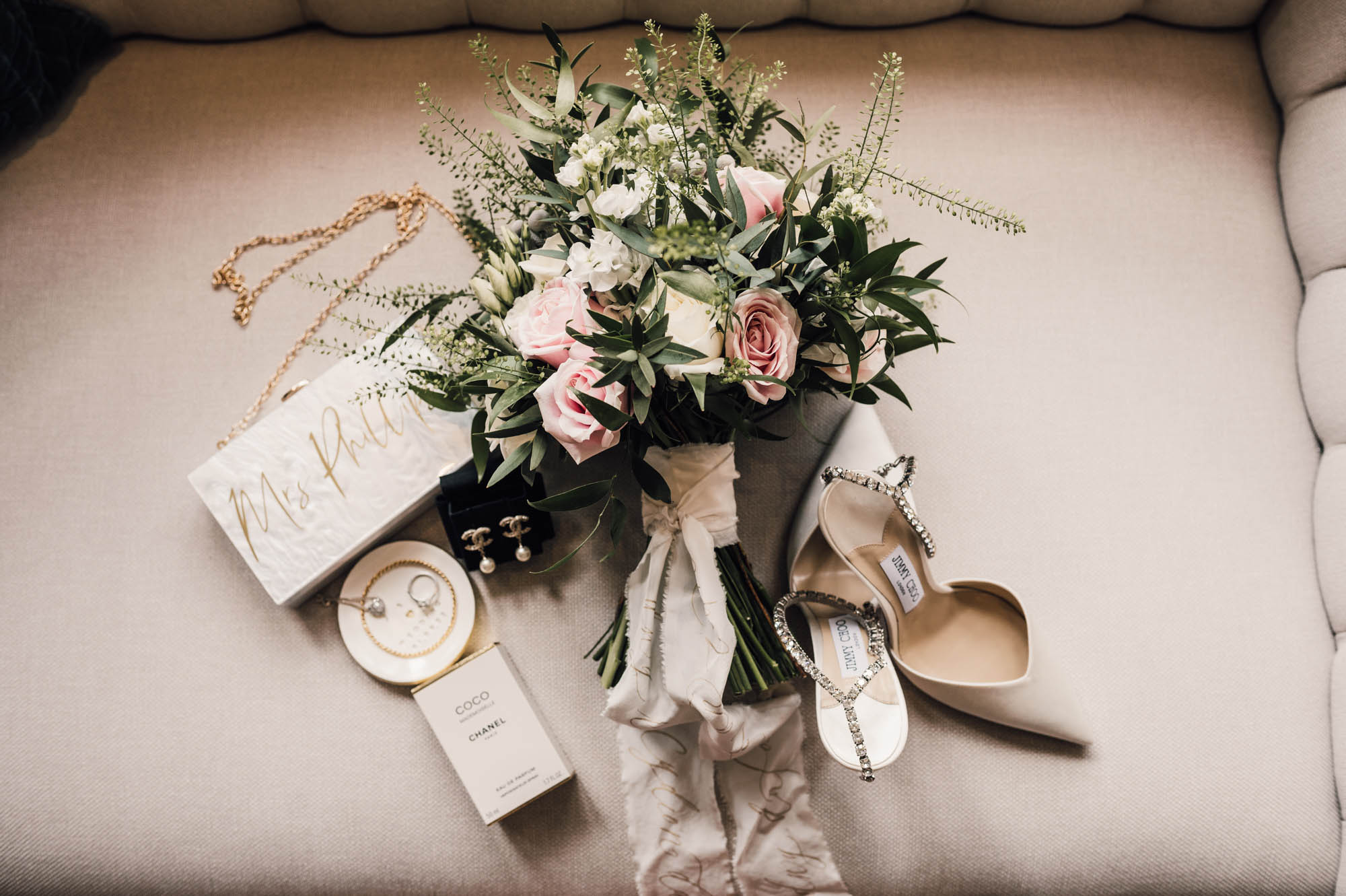 A wedding bouquet with ruscus leaves and pale pink roses tied with silk ribbon with personalised purse, shoes, jewellery and perfume. By Damien Vickers Photography