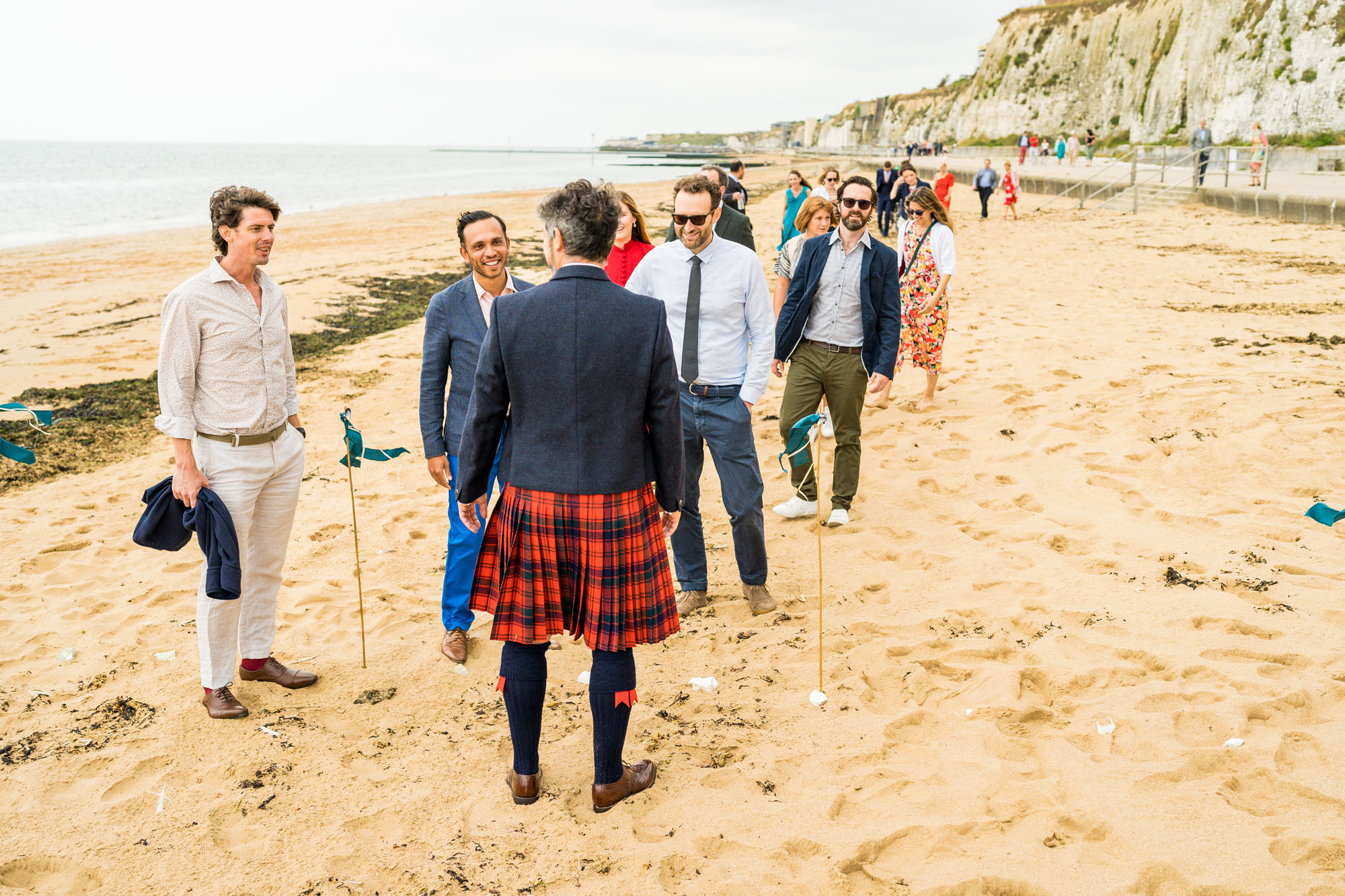 Guests walking along a beach to a relaxed modern wedding. The groom greets them, wearing a red tartan kilt. By Benjamin Toms, wedding photographer in Kent