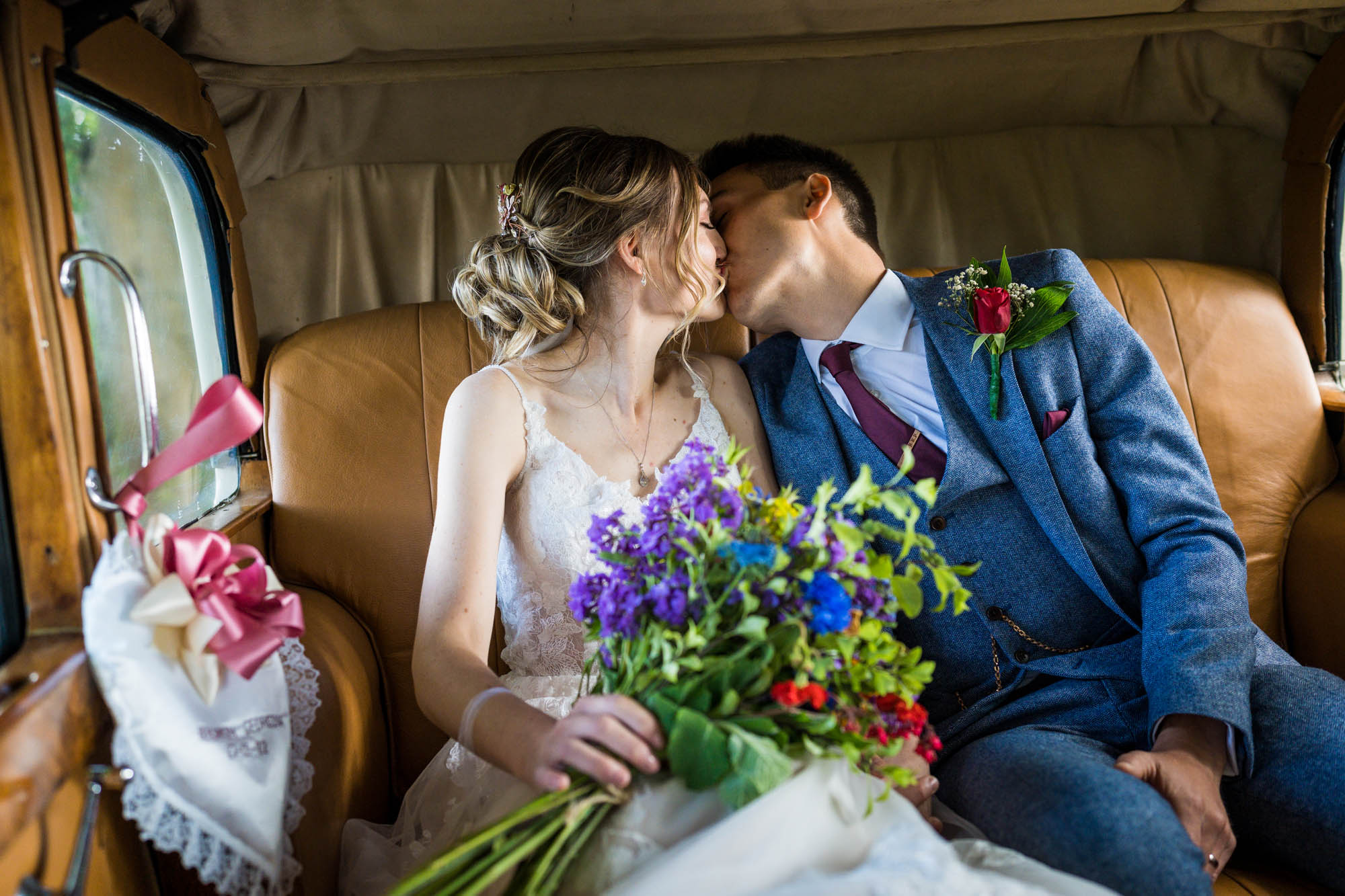 Newlyweds steal a kiss in the back of a vintage wedding car. She's carrying a colourful bouquet and he's wearing a blue suit. By wedding photographer Benjamin Toms, who's based in Kent, UK