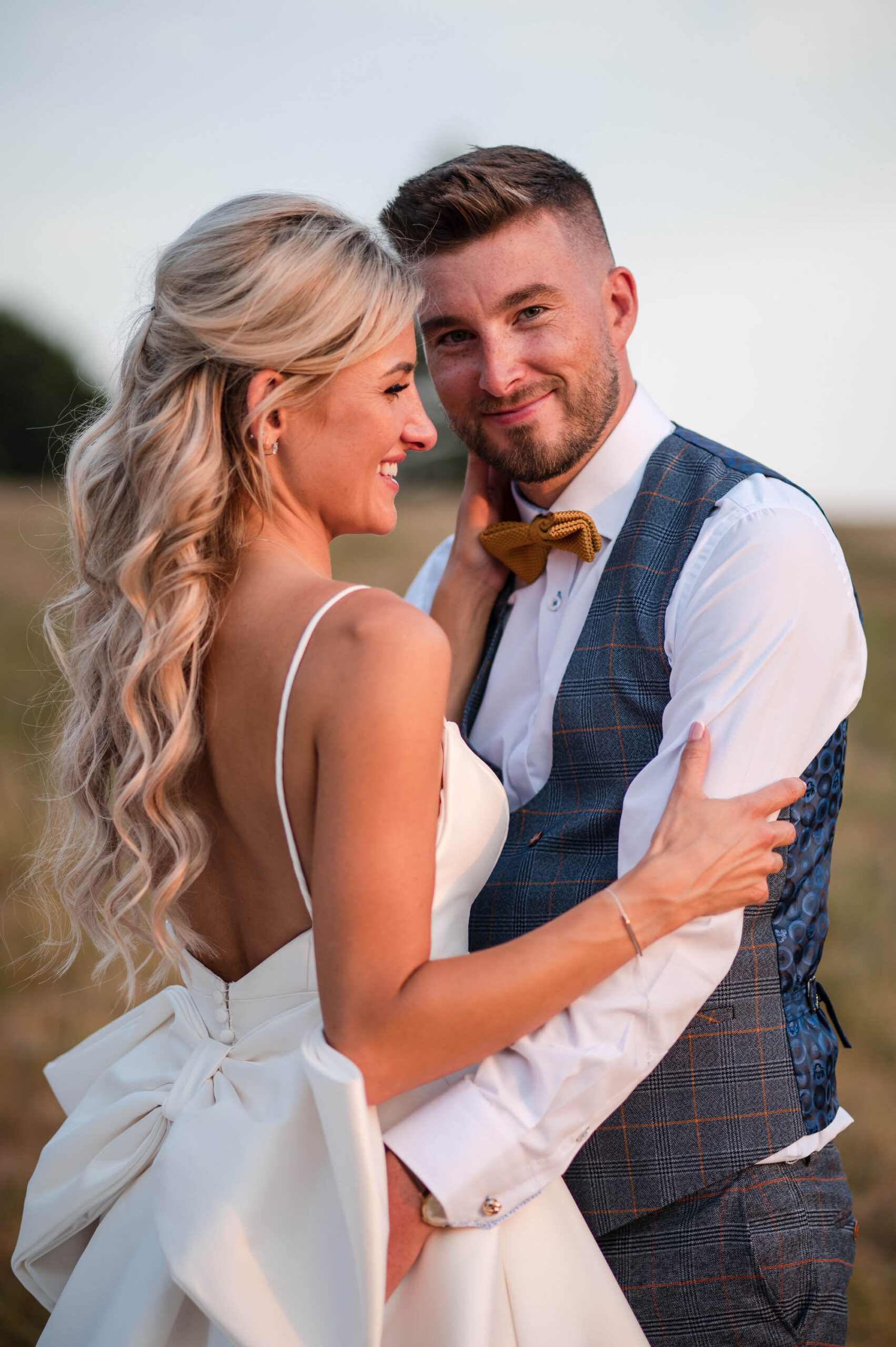 Bride and groom in golden sunset light. She's wearing a dress with straps, a low back and a decorative bow on the back. He's in a waistcoat with tan bowtie and both are smiling. By Libra Photographic 