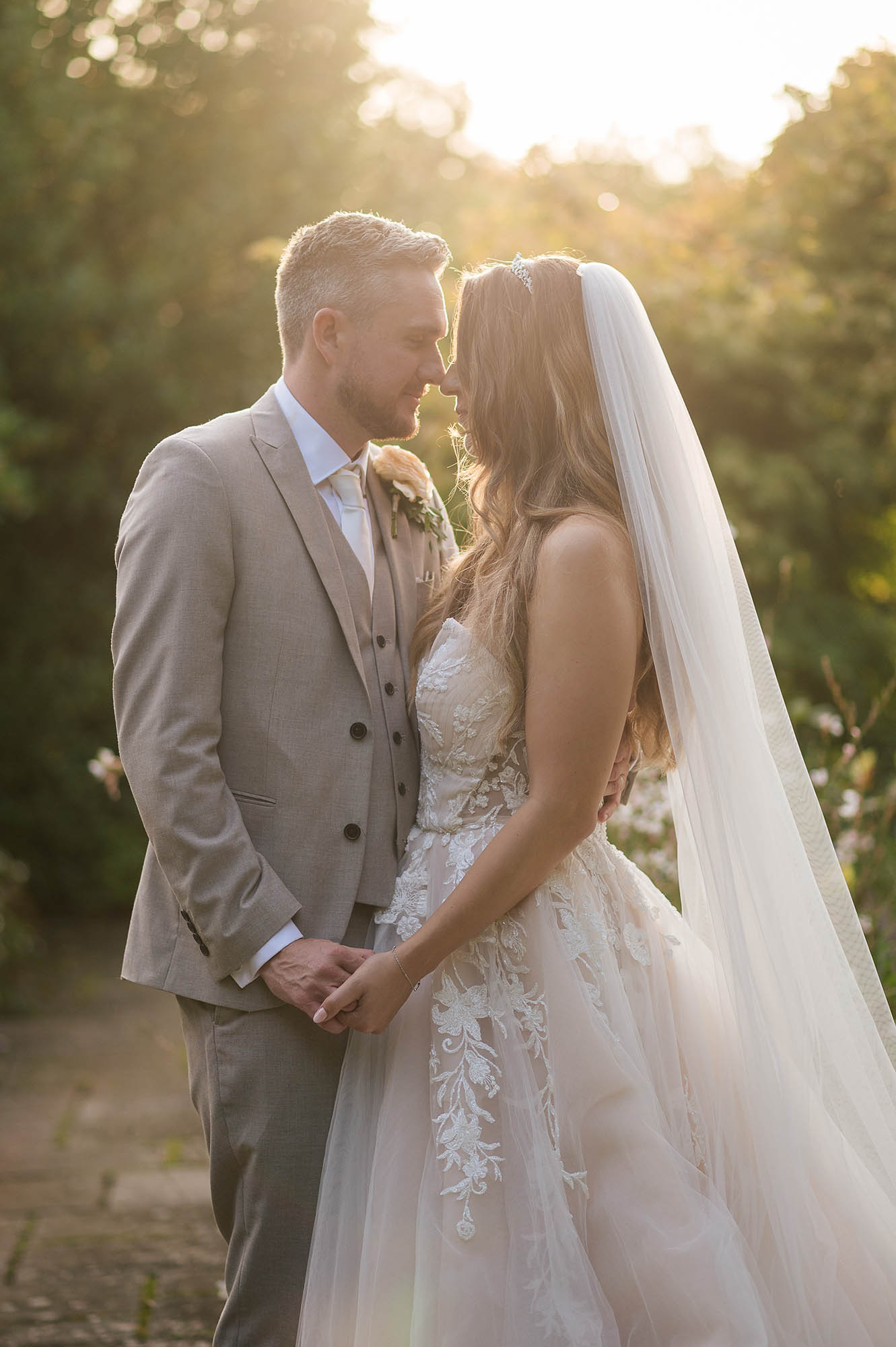 Groom and bride stood together with noses touching and holding hands. The sun is casting a soft glow on the couple. By Libra Photographic