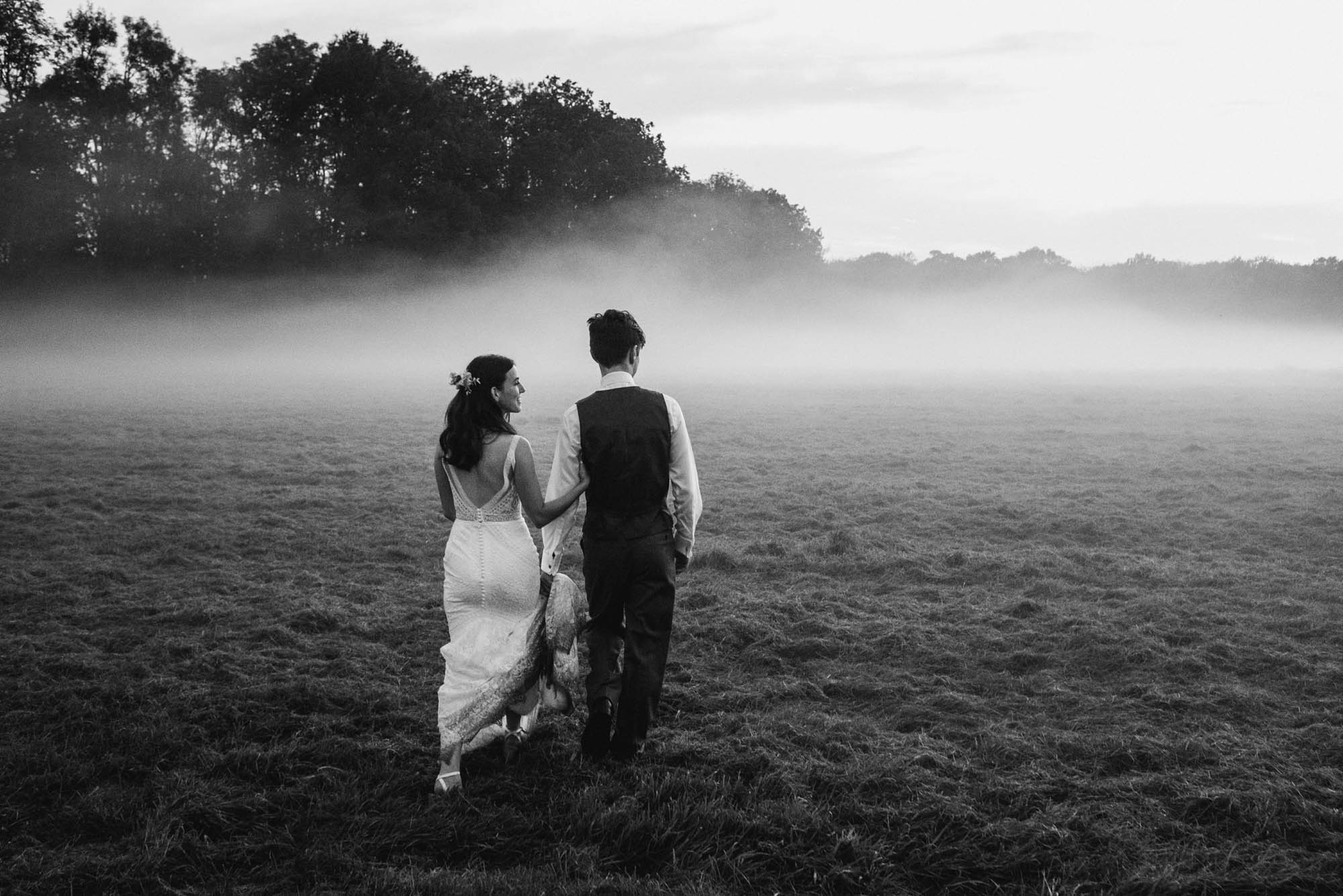 Sophie and Chris walk across a field with mist settling as evening falls on their wedding day. By York Place Studios at High Billinghurst Farm