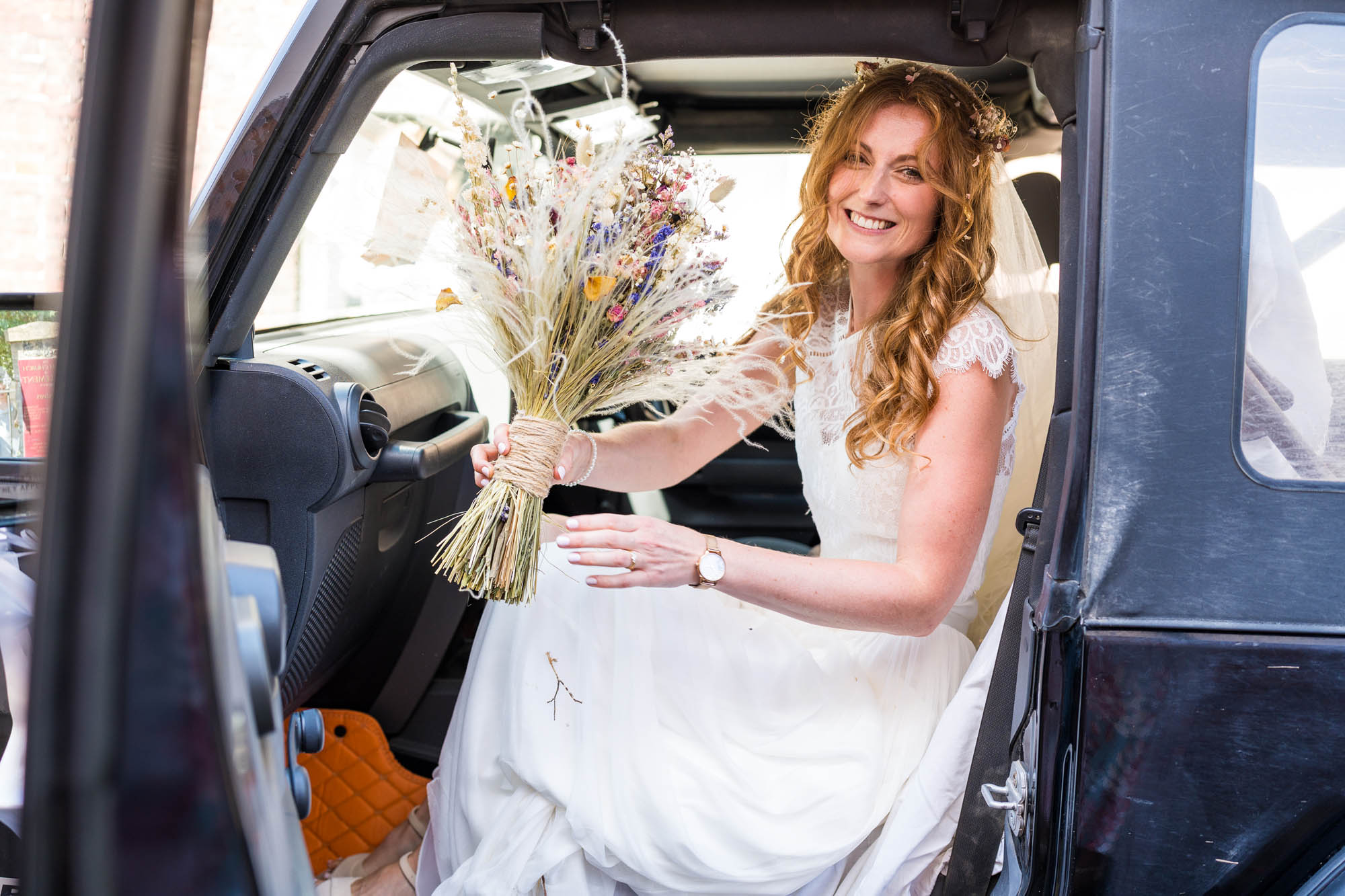 A happy bride in the front of a classic all terrain vehicle, holding a bunch of wild flowers and grasses. Taken by Kent wedding photographer Benjamin Toms