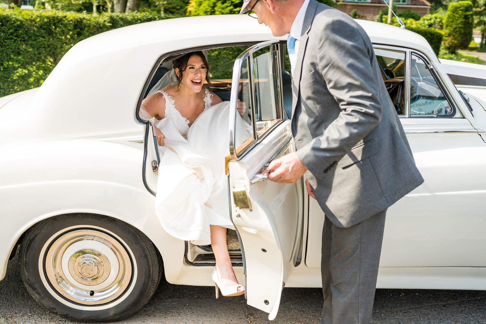 A bride steps from a luxury vintage car, helped by a smartly dressed older man who is holding the door for her. Captured by Benjamin Toms Photography in Kent