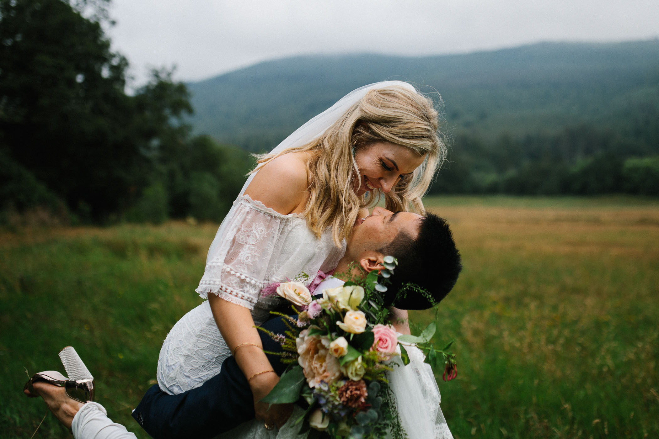 A bride is lifted up by her groom and they're about to kiss. She's holding a bouquet of ivory and pink flowers. By Luke Flint Photography