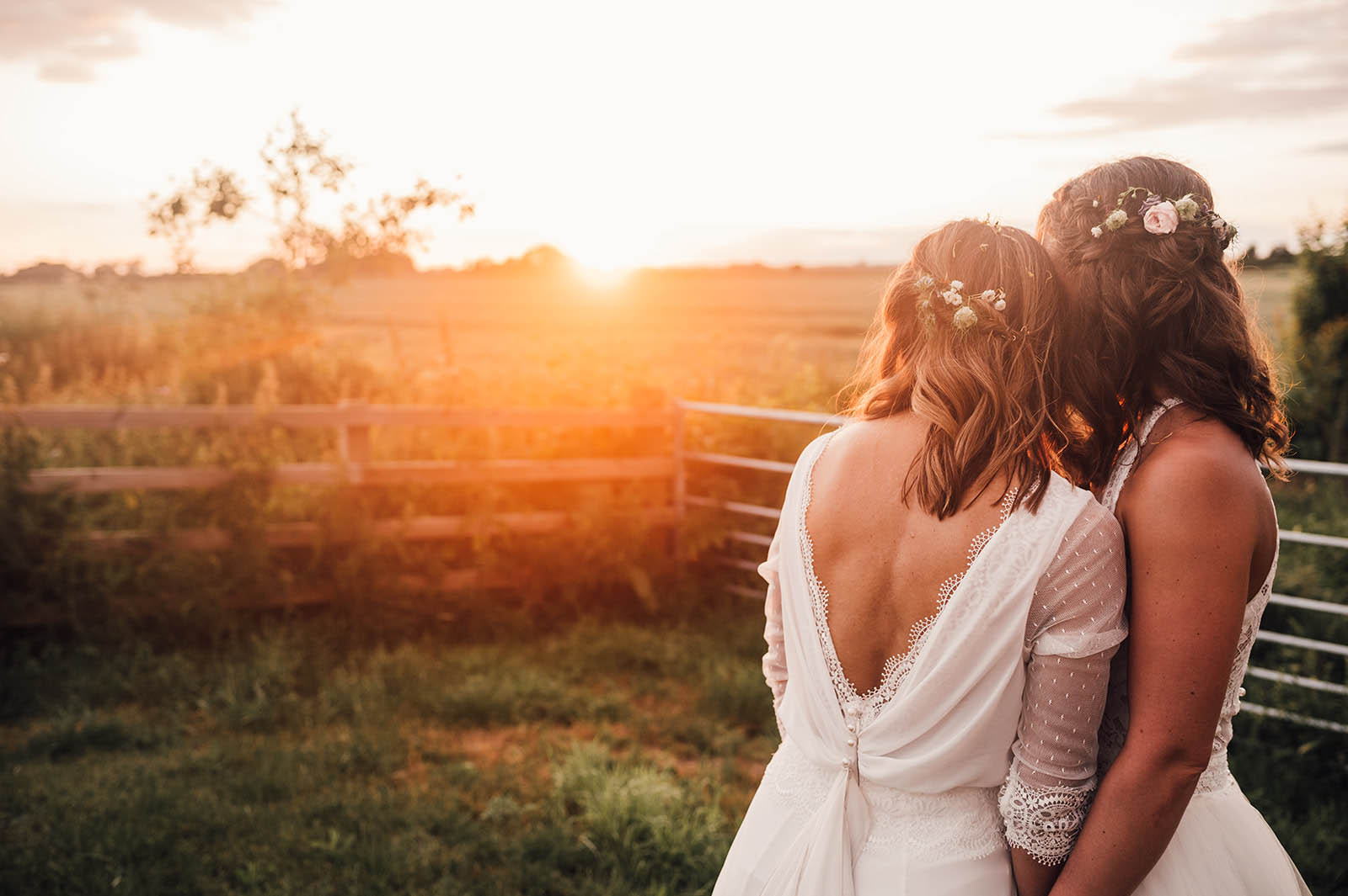 Beautiful golden hour photograph of two brides at Willow Grange Farm in Cambridge. They're facing away from the camera, huddled close to each other and looking at the sunset. Both have flowers in their hair. By Damien Vickers Photography