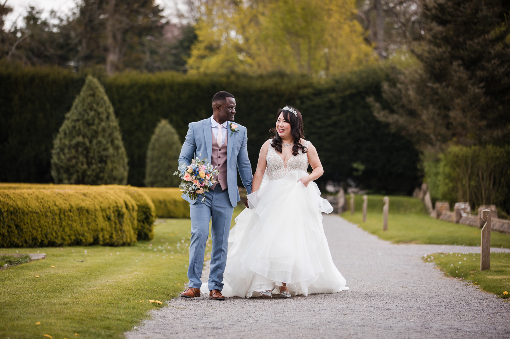 A groom and bride walk down a pathway through landscaped gardens at Tortworth Court wedding venue. He's holding the bouquet and wearing a pale blue suit. She's smiling towards him as they walk, wearing a ruffled dress and headpiece. Photographer credit Greg Shingler from Wiltshire