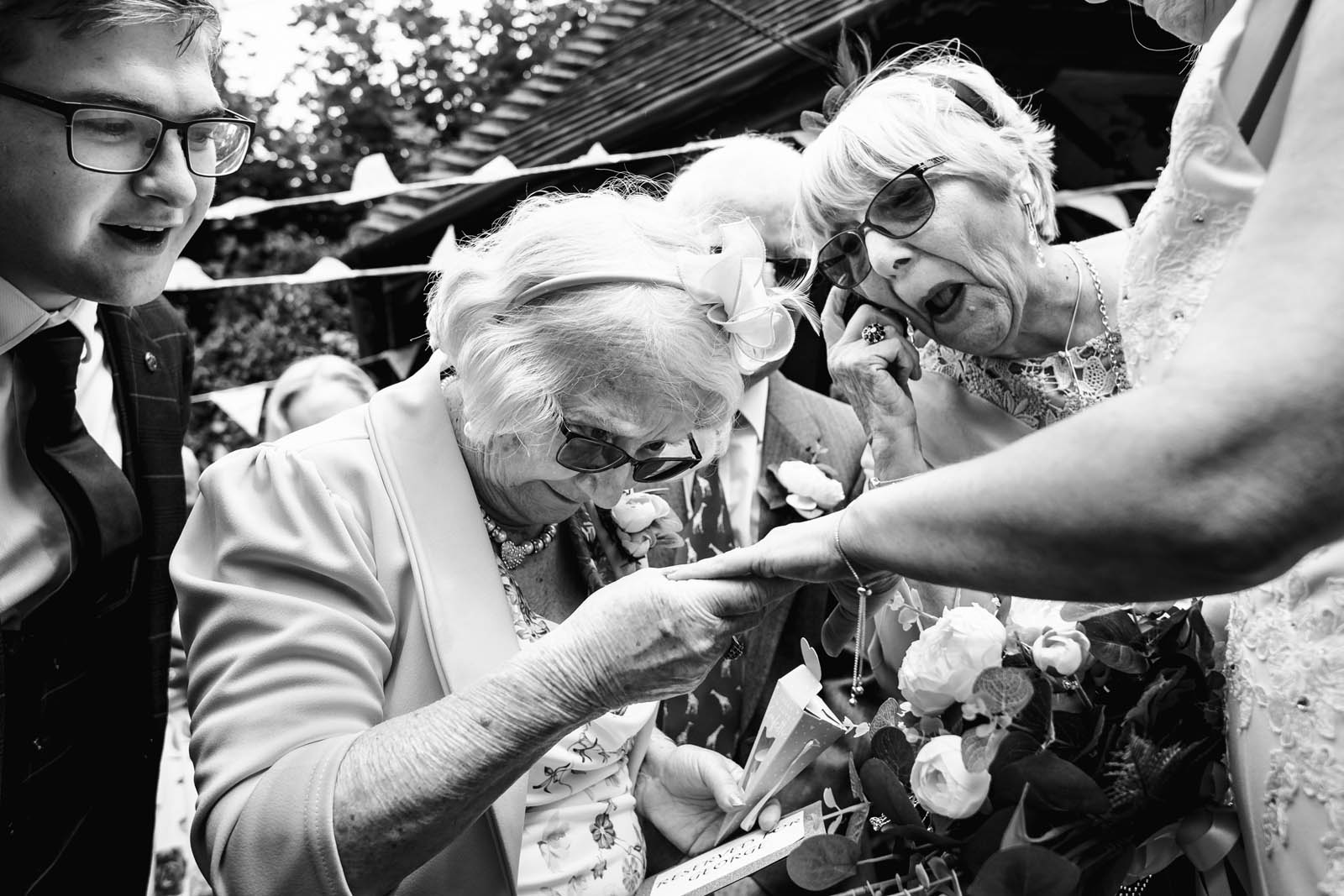 A grandmotherly wedding guest is inspecting a bride's ring at close quarters while other guests peer over her shoulder to see.
